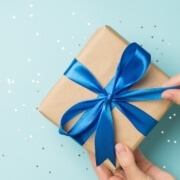 First person top view photo of hands unpacking craft paper gift box with blue satin ribbon bow over shiny sequins on isolated pastel blue background with blank space