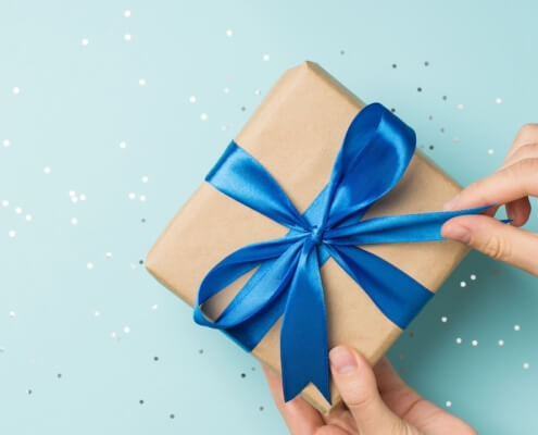 First person top view photo of hands unpacking craft paper gift box with blue satin ribbon bow over shiny sequins on isolated pastel blue background with blank space